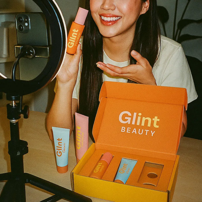 A woman records a video reviewing cosmetics. She holds one of the bottles up to a ring light; the other products are in a mailer box in front of her on a desk.