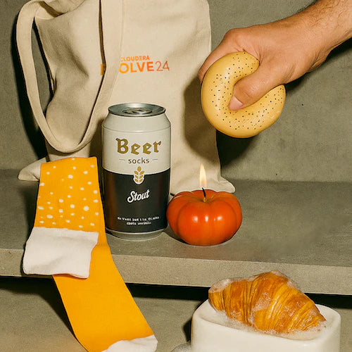 Event gift bag for an ai conference with socks that look like beer, a candle that looks like a tomato, a stress toy that looks like a bagel and soap that looks like a croissant.