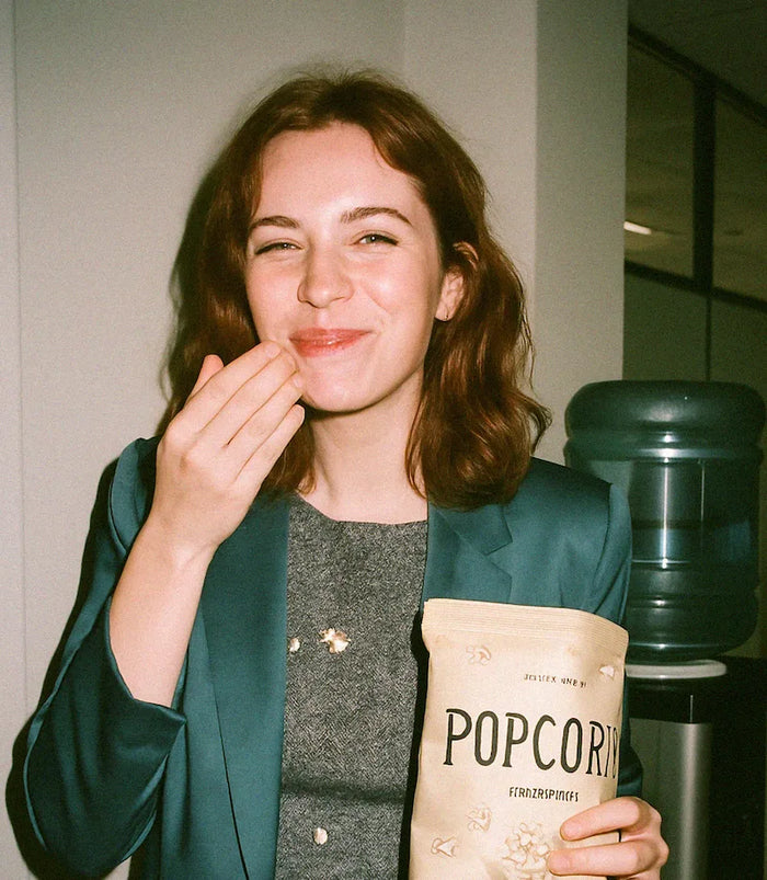 A woman in a teal blazer eats popcorn in front of a water cooler in an office.
