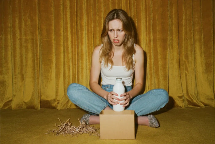 A young woman sits on mustard yellow carpet in front of mustard yellow curtains, holding a white water bottle. She is looking at it with disappointment. A kraft box sits in front of her.